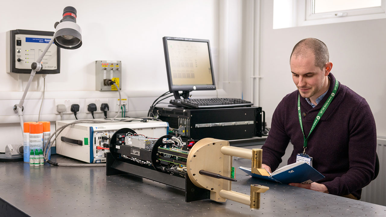 Man with shaved head wearing purple jumper reads blue book while sat at desk in laboratory