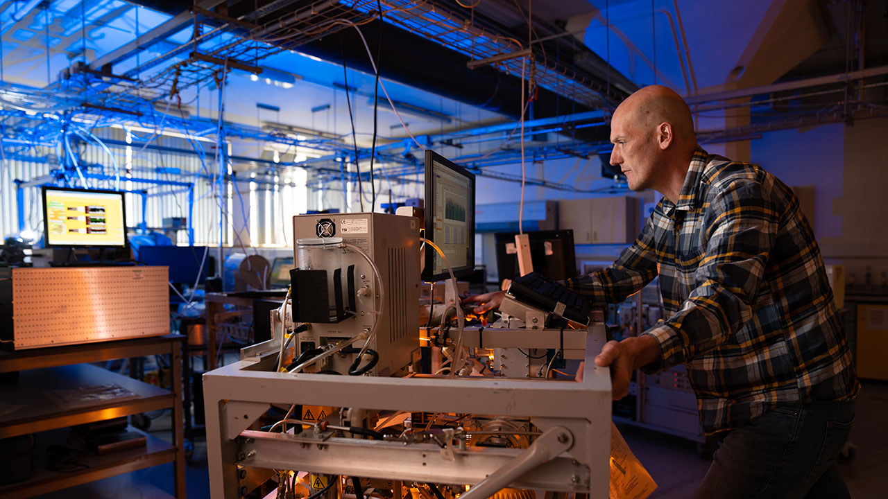 A man in a checked shirt in a laboratory leans over a piece of scientific equipment looking at a computer screen